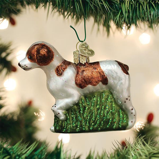 Hand-painted blown glass Brittany Spaniel ornament with brown and white markings, glittered green grass, and a heart charm.