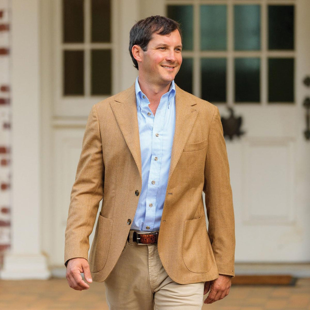 Man wearing a camel herringbone sports jacket with notch lapels and patch pockets over a light blue button-down shirt.