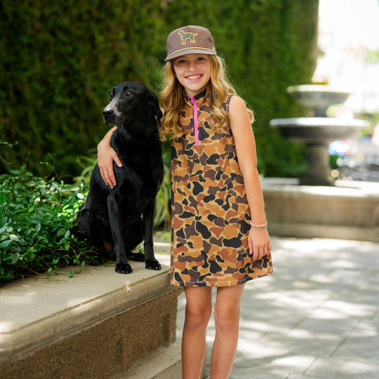 Girl wearing a vintage camo performance dress with a pink quarter-zip neckline and built-in shorts, standing with a black dog.