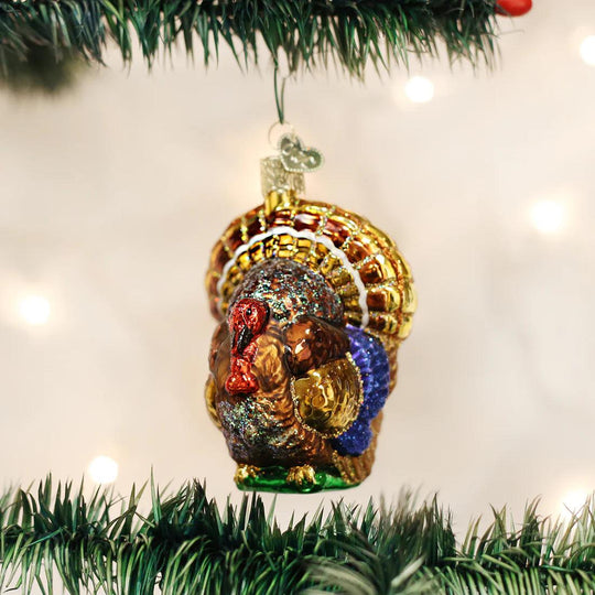 Hand-painted blown glass turkey ornament with glitter accents, hanging on a green pine branch for Thanksgiving decor.
