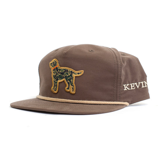 A brown 5-panel snapback cap featuring a green camouflage dog patch on the front and the name 'KEVIN'S' embroidered in cream on the side, set against a plain white background.