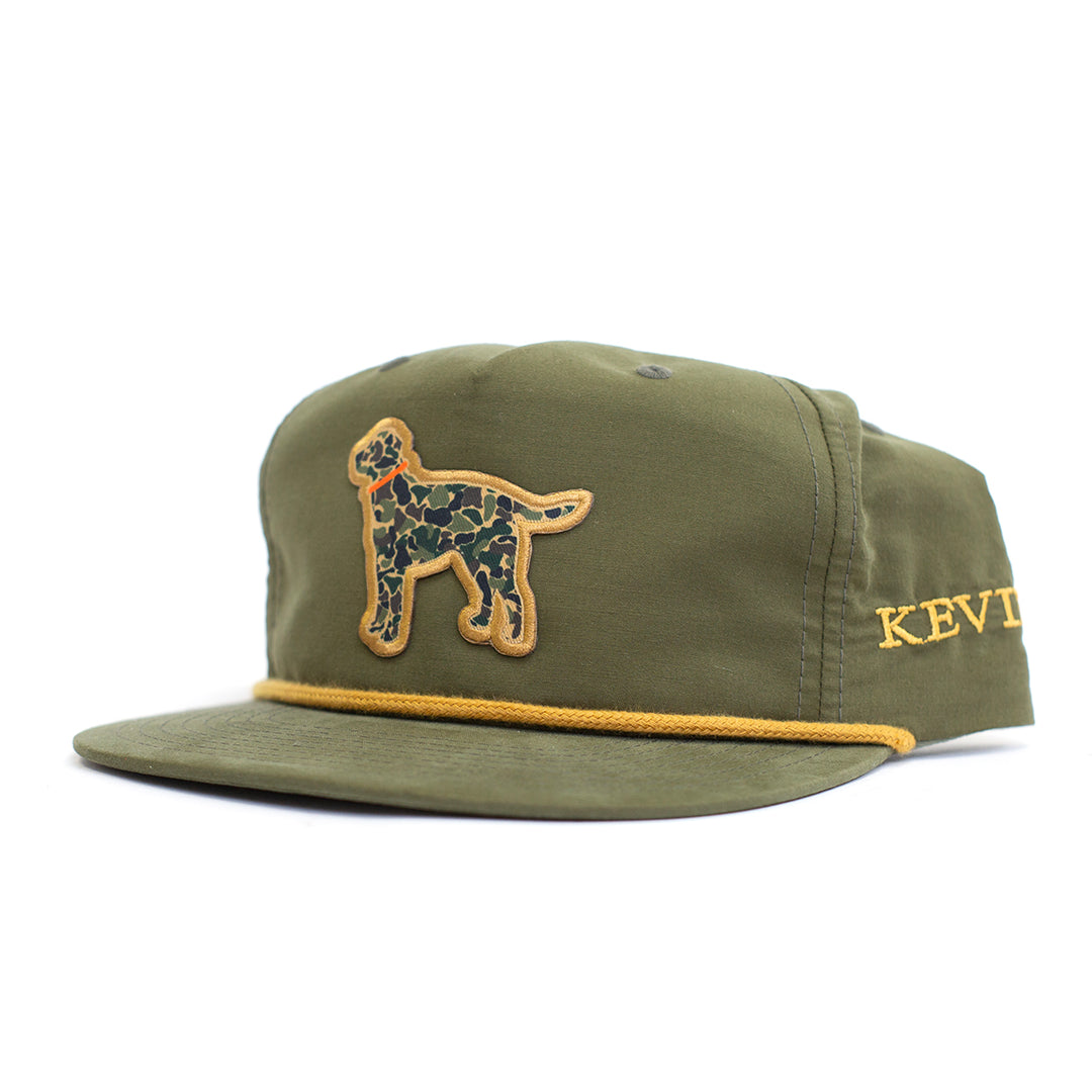 An olive green 5-panel cap with a yellow embroidered dog patch on the front and yellow embroidered text on the side, shown on a white background.