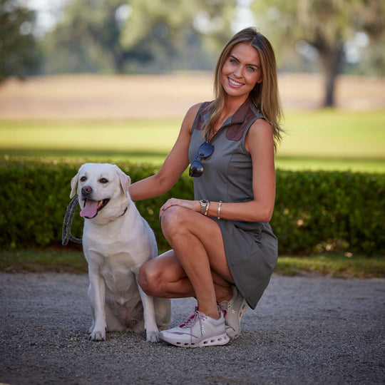 Woman wearing an olive green performance sporting dress with a suede-trimmed quarter-zip neckline and a white Labrador.