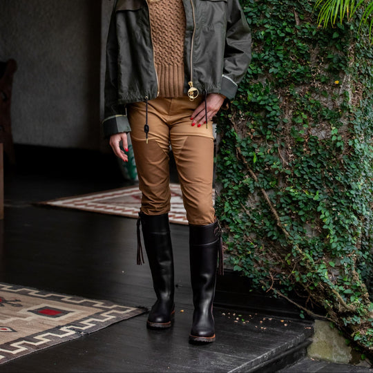 Woman wearing tan high-rise stretch briar pants with reinforced front panels, styled with a brown sweater and tall black boots.