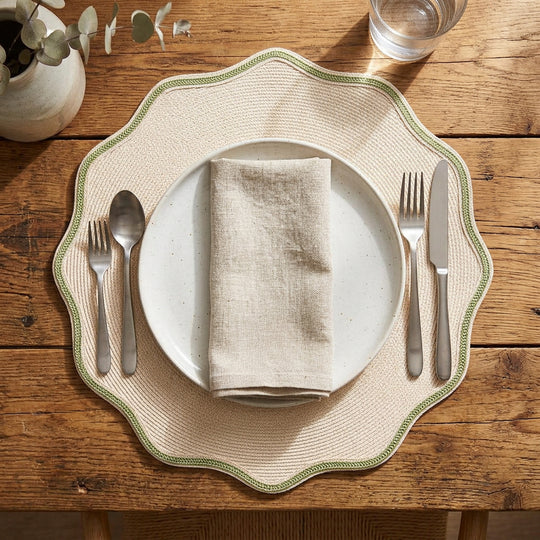 16-inch round sand-colored woven placemat with scalloped green piped edges on a rustic wooden table.