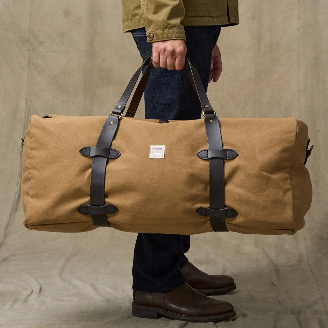 Tan Filson large duffle bag in rugged twill with dark brown bridle leather straps and handles held by a person.