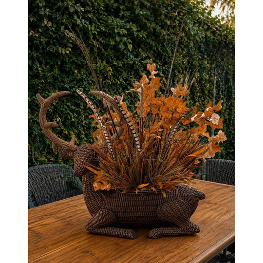 Hand-woven brown willow stag-shaped party bucket filled with autumn leaves and feathers on a wooden outdoor table.