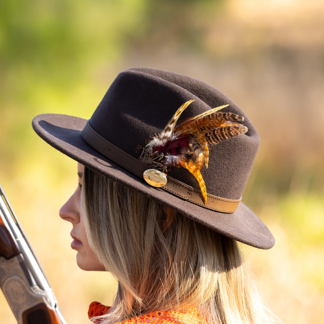 Dark brown wool felt Suffolk Fedora with a leather band, gold logo emblem, and decorative pheasant feather trim.