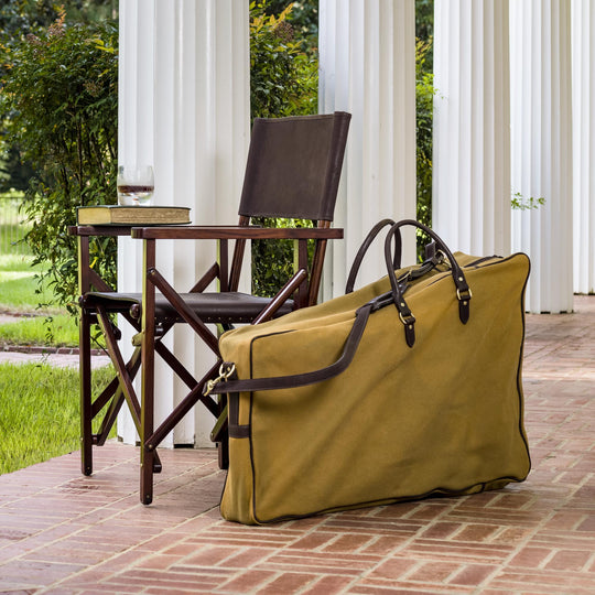 Tan canvas carrying case with leather trim, handles, and shoulder strap next to a folding wooden salon chair on a patio.