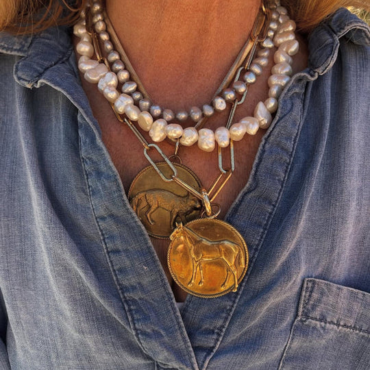 Gold horse pendant on a layered paperclip chain styled with pearl necklaces over a denim shirt.