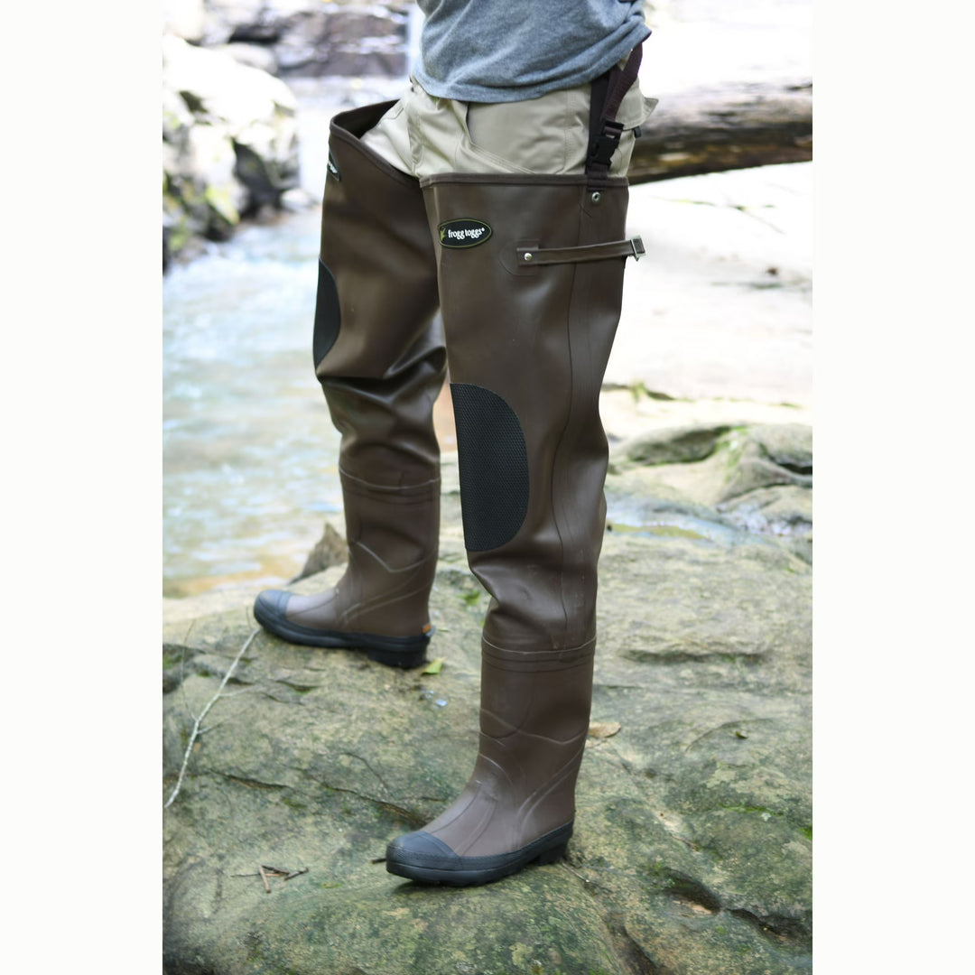 Frogg Toggs brown rubber hip waders with black knee pads and adjustable belt straps standing on a rocky riverbank.