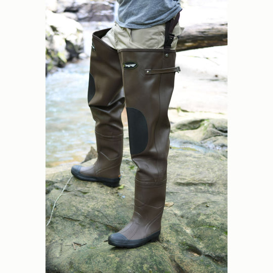 Frogg Toggs brown rubber hip waders with black knee pads and adjustable belt straps standing on a rocky riverbank.