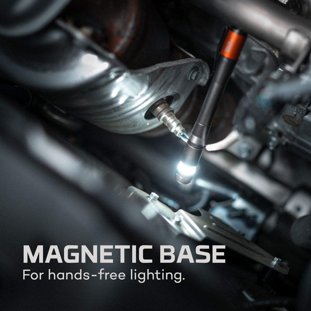 Black and orange NEBO Inspector 500+ penlight magnetically attached to a car engine for hands-free illumination.